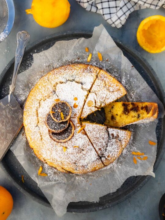 Overhead of an orange ricotta cake on baking paper and a round grey terrazo tray with one piece on its side, with icing sugar and dried orange slices on top.