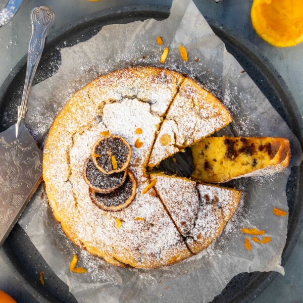 Overhead of an orange ricotta cake on baking paper and a round grey terrazo tray with one piece on its side, with icing sugar and dried orange slices on top.