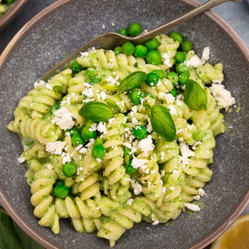Closeup of a mottled grey bowl of fusilli pasta with peas with fresh peas and feta and herbs on top on a grey background with a fork in the bowl and lemon halves and beside it.