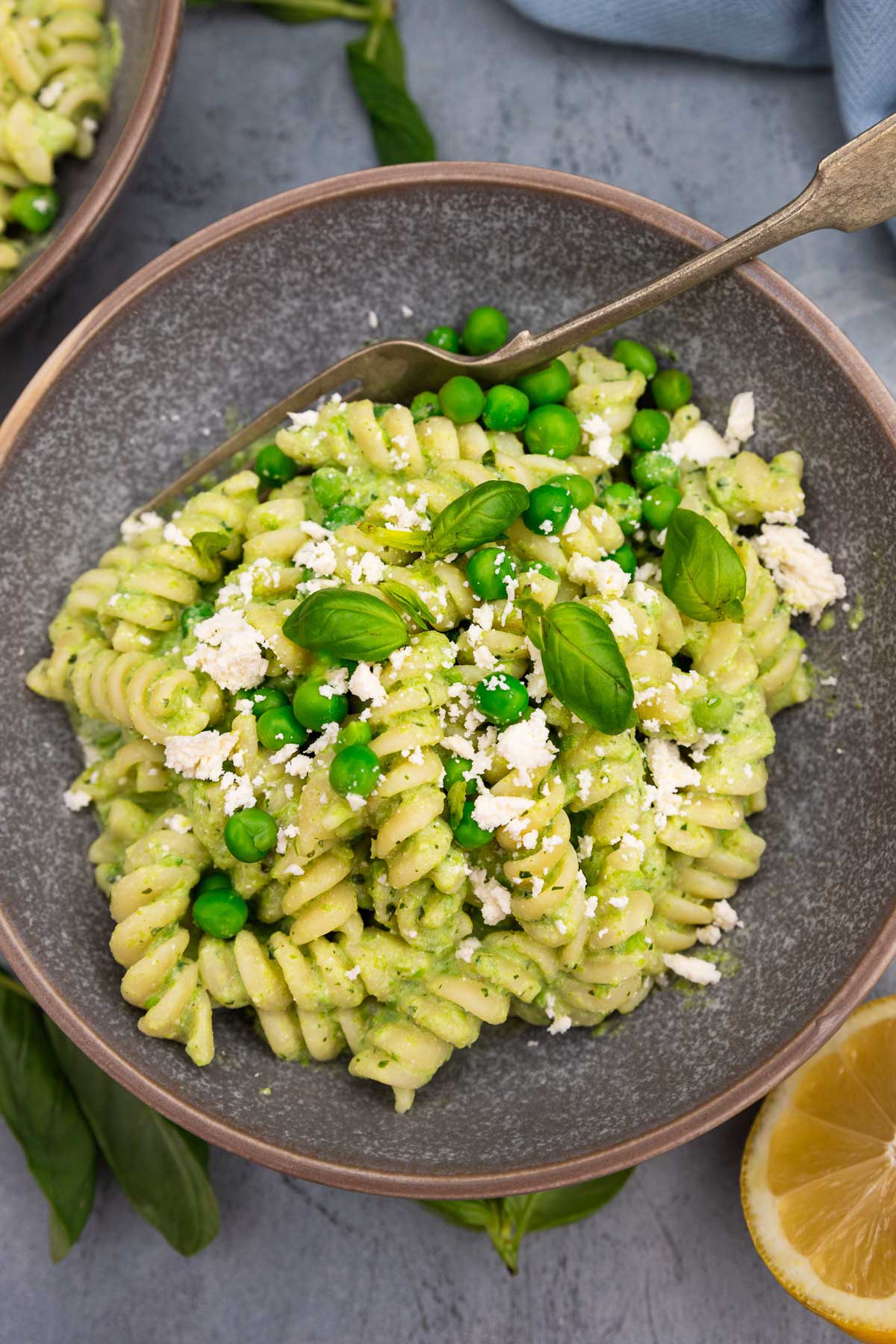 Closeup of a mottled grey bowl of fusilli pasta with peas with fresh peas and feta and herbs on top on a grey background with a fork in the bowl and lemon halves and beside it.