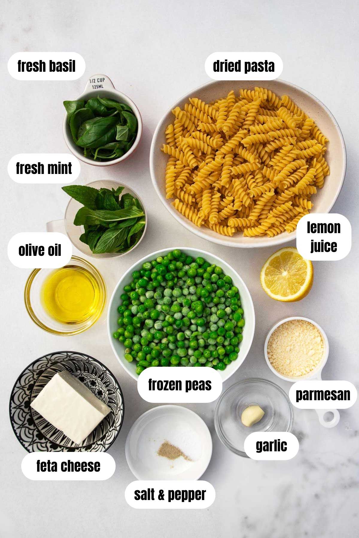 Overhead of all ingredients for a creamy pea pasta including fusilli pasta, feta, salt and pepper, frozen peas, garlic, parmesan, lemon juice parmesan, olive oil and fresh herbs, all on a marble background.