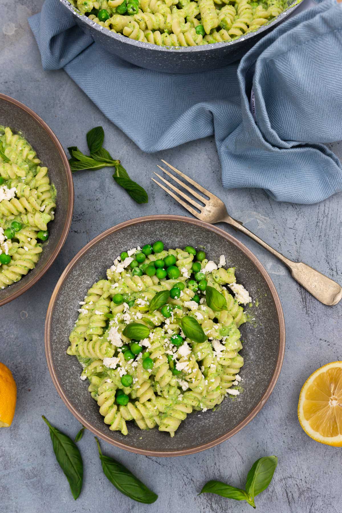 Overhead view of two bowls of creamy pea pasta with fresh peas, herbs and feta on top on a blue distressed background with more fresh herbs, lemon halves and a pale blue tea towel around them.