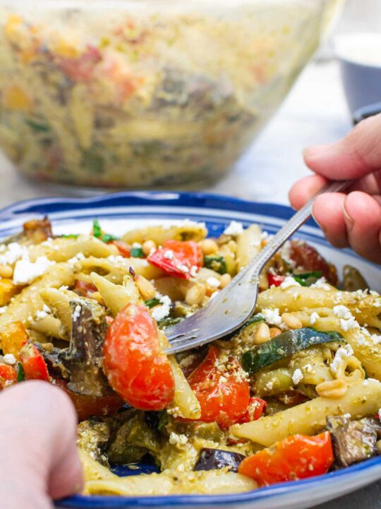 A closeup of someone's hands scooping up some penne pesto pasta with colorful vegetables from a blue-rimmed bowl with more of the pasta in the background in a Pyrex bowl.