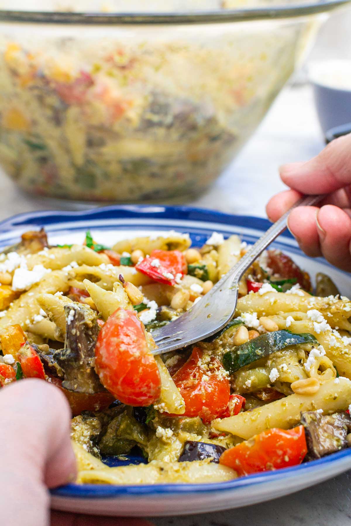 A closeup of someone's hands scooping up some penne pesto pasta with colorful vegetables from a blue-rimmed bowl with more of the pasta in the background in a Pyrex bowl.