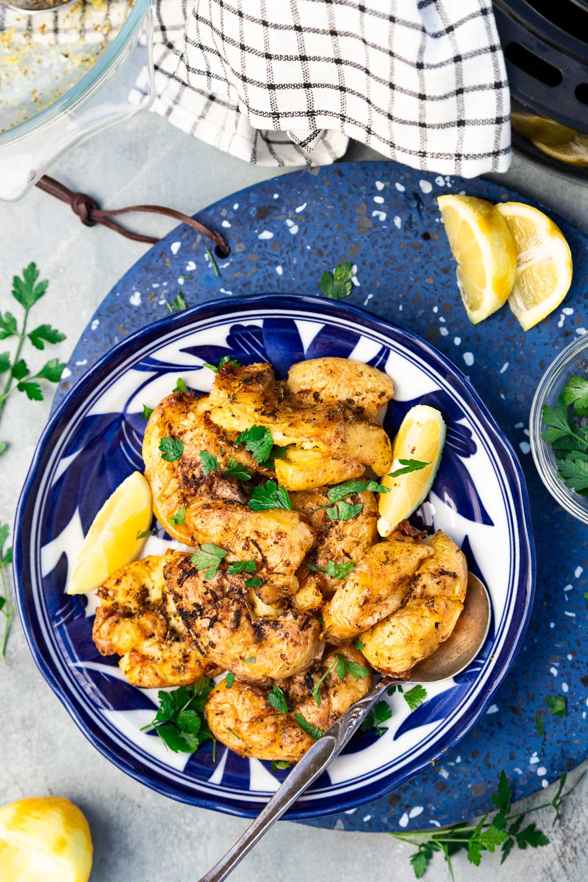 Air fried smashed potatoes in a decorative blue dish with a decorative spoon, all on a blue terrazo trivet with lemon, parsley and a black and white tea towel around it.