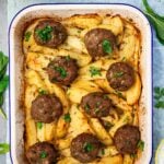 Overhead of Greek biftekia or hamburgers on top of lemon potatoes in a blue-rimmed white baking pan, all on a light blue tea towel with herbs around the pan and the edge of a Greek salad and tzatziki visible.