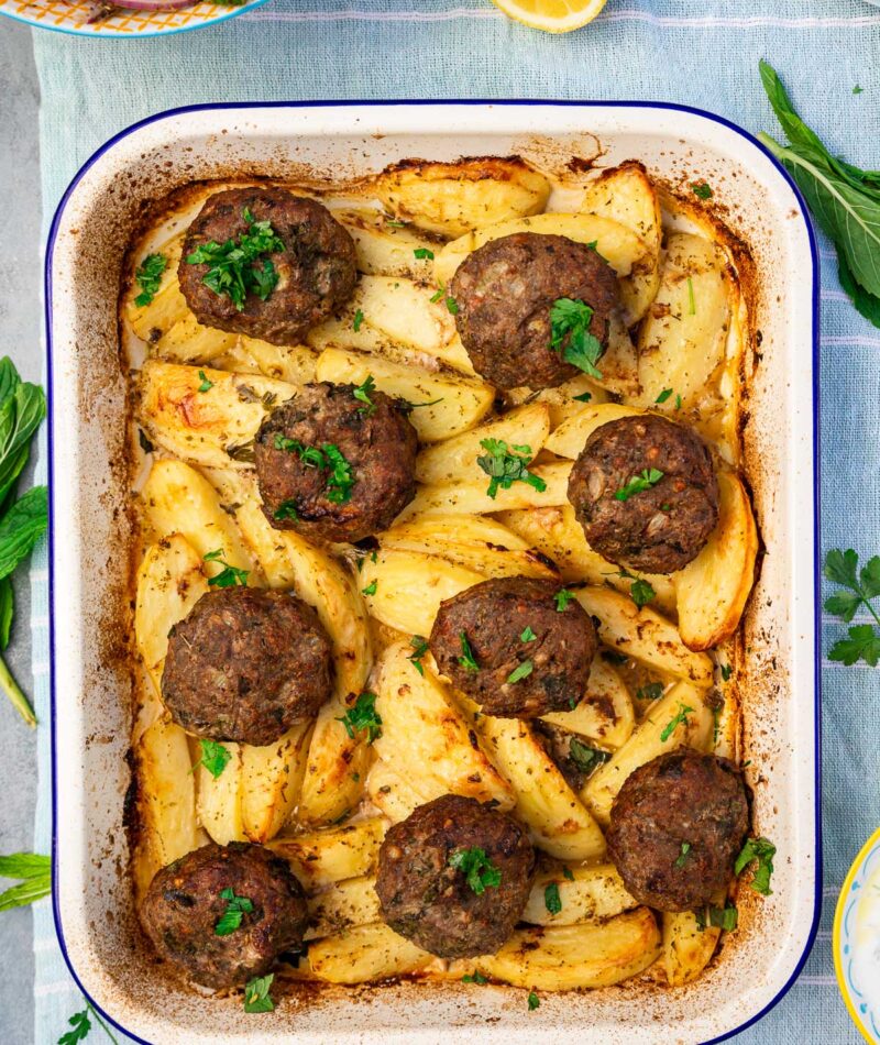 Overhead of Greek biftekia or hamburgers on top of lemon potatoes in a blue-rimmed white baking pan, all on a light blue tea towel with herbs around the pan and the edge of a Greek salad and tzatziki visible.