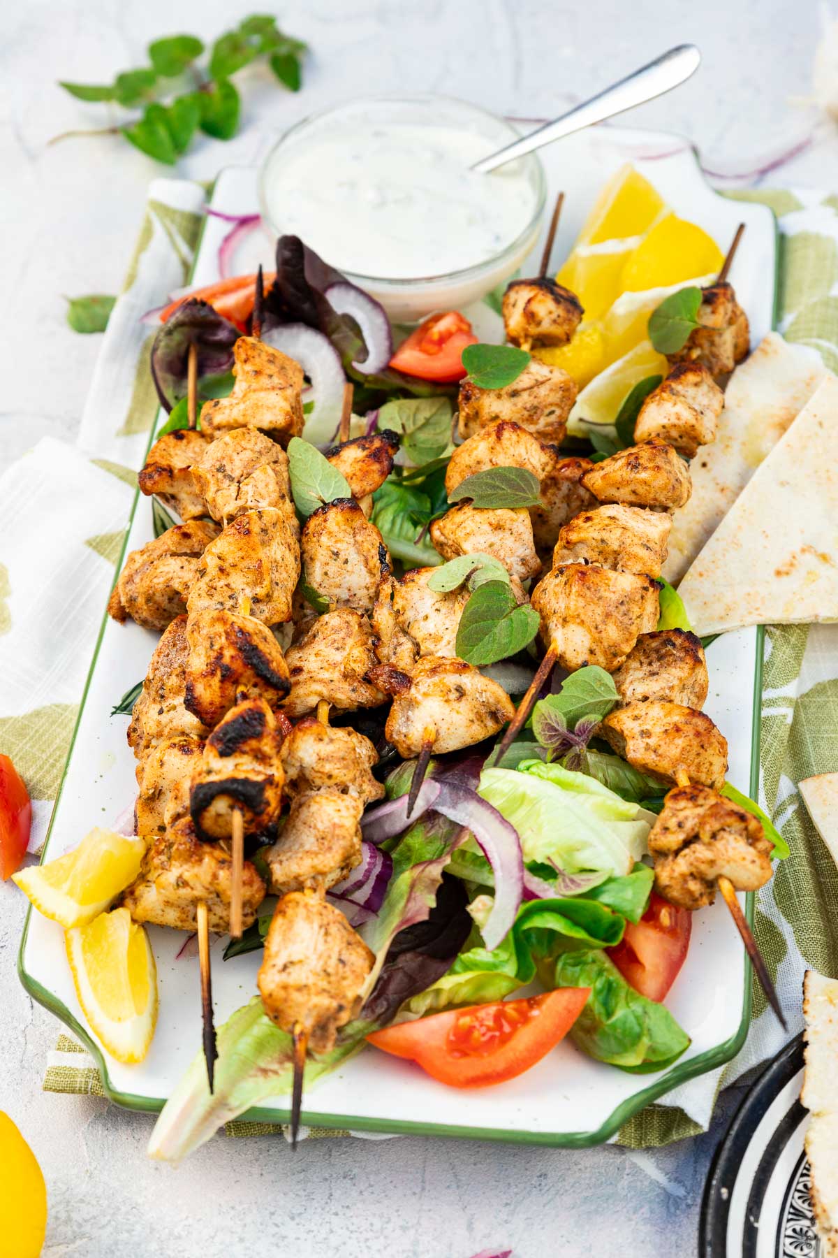 A rectangular platter of baked chicken kabobs on a bed of simple salad and with lemon segments and a white dip on a green and white tea towel and on a pale textured background.