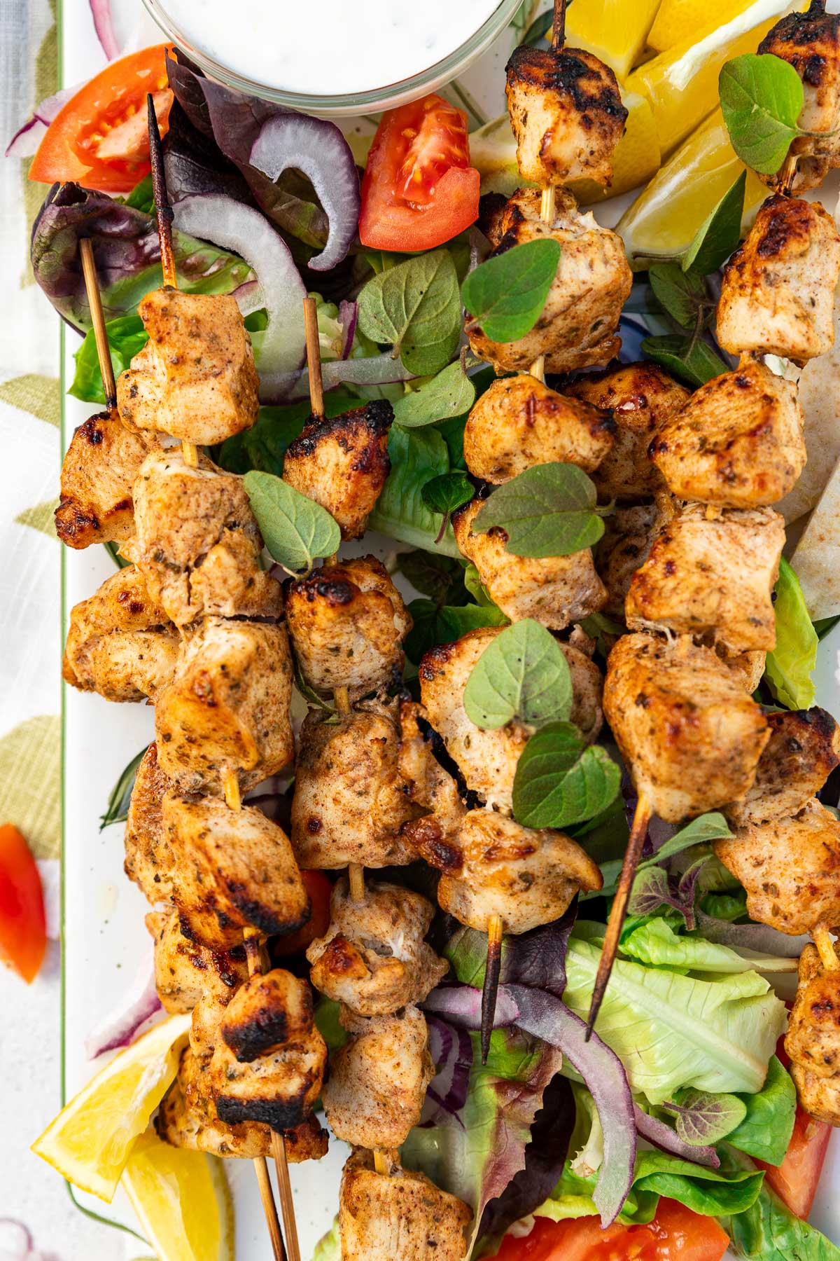 Overhead closeup of a rectangular platter of baked chicken kabobs on a bed of simple salad and with lemon segments and a white dip on a green and white tea towel and on a pale textured background.