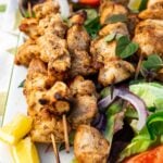 Closeup of a rectangular platter of baked chicken kabobs on a bed of simple salad and with lemon segments and a white dip on a green and white tea towel and on a pale textured background.