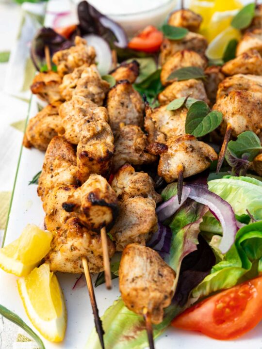 Closeup of a rectangular platter of baked chicken kabobs on a bed of simple salad and with lemon segments and a white dip on a green and white tea towel and on a pale textured background.