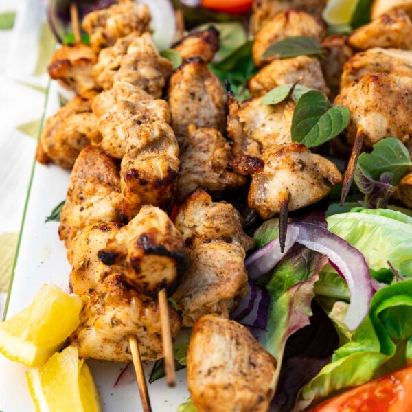 Closeup of a rectangular platter of baked chicken kabobs on a bed of simple salad and with lemon segments and a white dip on a green and white tea towel and on a pale textured background.