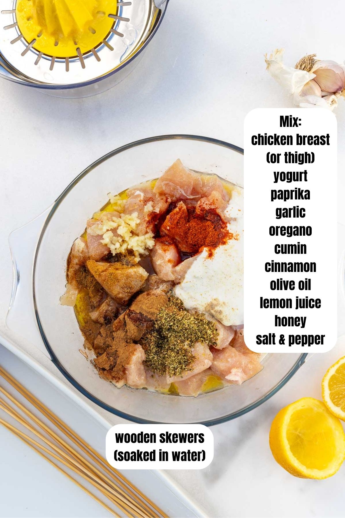 Overhead view of the ingredients needed for baked chicken kabobs including a bowl with chicken cubes and all the marinade ingredients, wooden skewers soaking in water and squeezed lemon and garlic around them on a marble background.