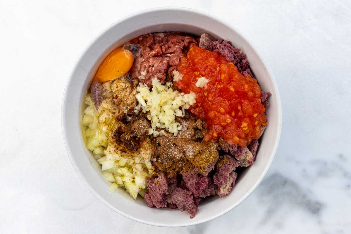 Overhead of all biftekia or Greek burger ingredients in a white bowl, including ground beef, spices, salt and pepper, soaked bread, crushed garlic, an egg, onion and grated tomato.
