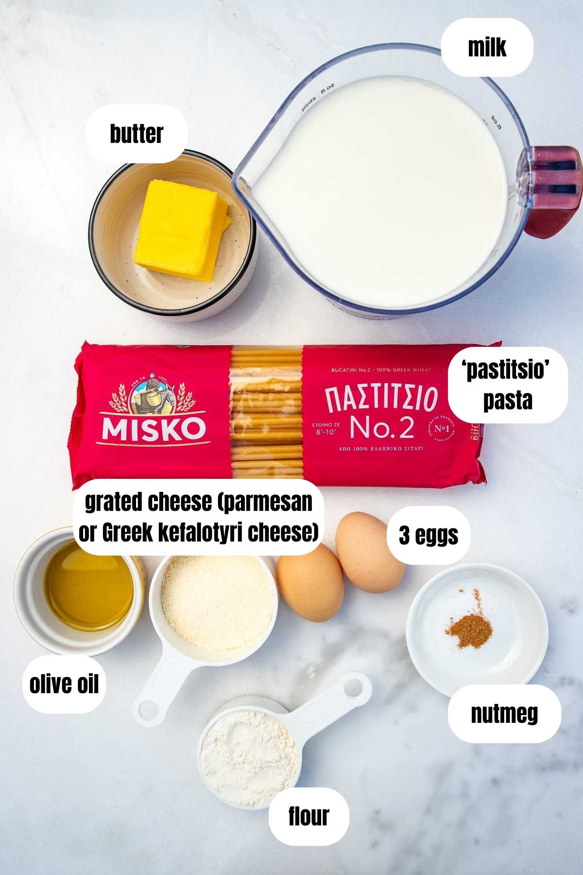 Overhead on marble background of all ingredients to make Greek pastitsio pasta and bechamel sauce including pastitsio pasta, olive oil, hard cheese, flour, milk, nutmeg, butter and eggs.