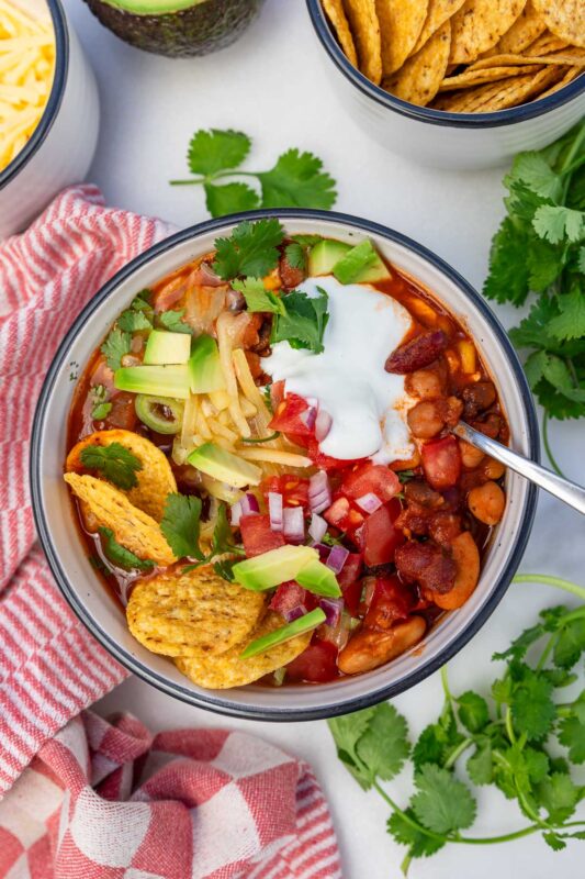 A dish of 5 bean chili with a spoon and with salsa, grated cheese, avocado , coriander, sour cream and nachos on it.