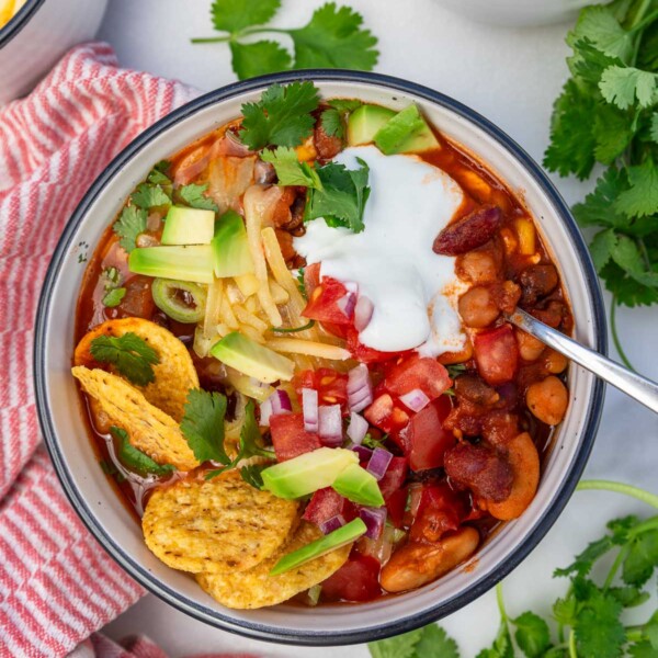 A dish of 5 bean chili with a spoon and with salsa, grated cheese, avocado , coriander, sour cream and nachos on it.