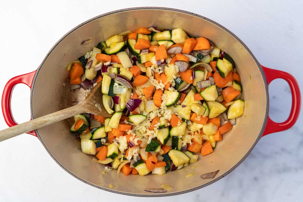 Chopped vegetables and crushed garlic in a red cast iron pot with a wooden spoon in it.