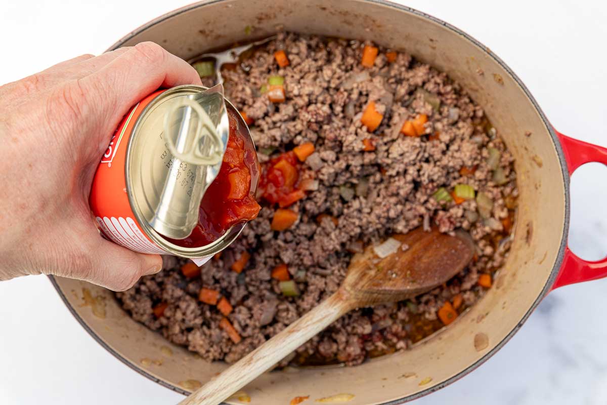 Overhead of someone adding a can of diced tomatoes to a dish of Greek ragu sauce for pastitsio in a red cast iron pan with a wooden spoon in it.