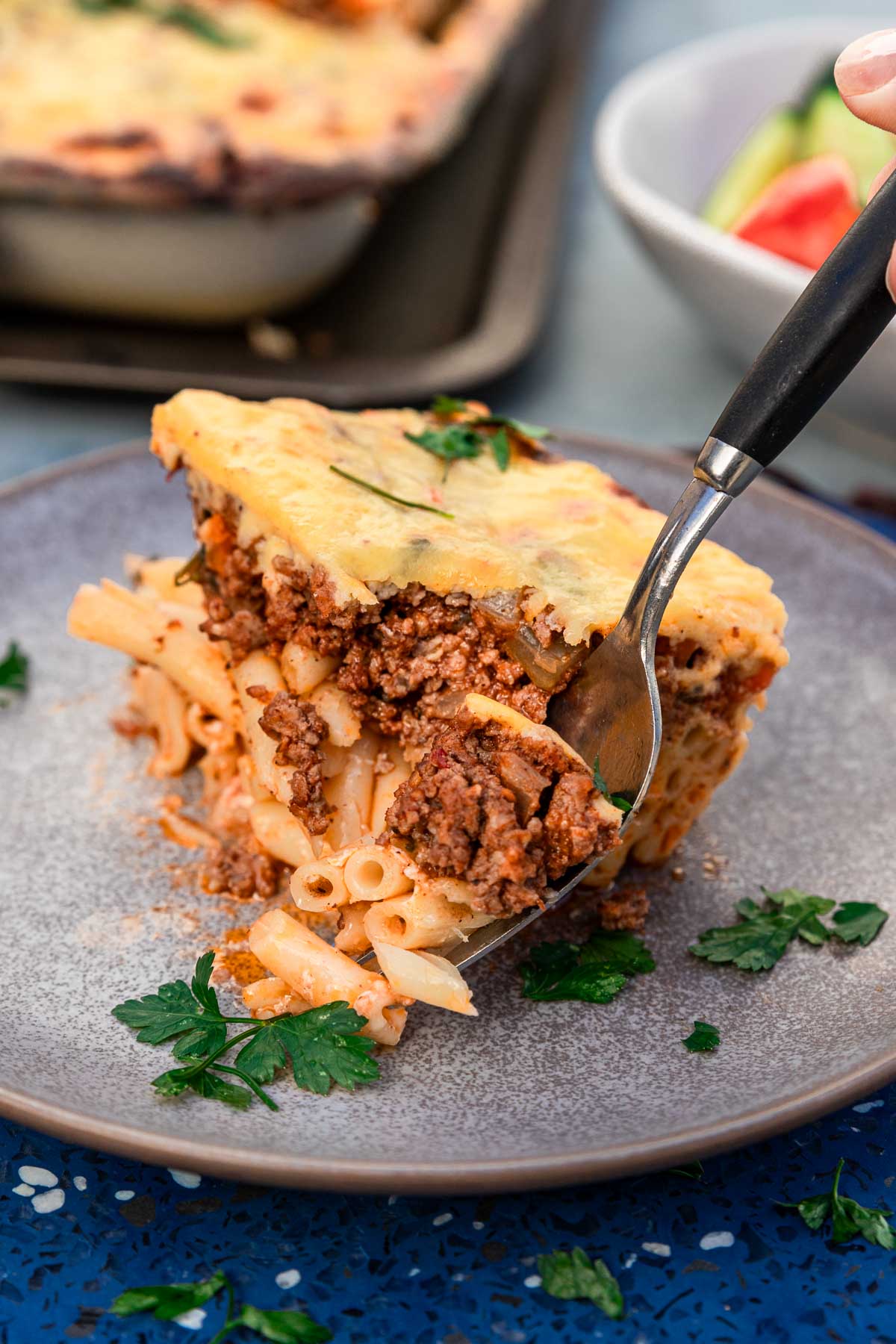 Someone eating with a fork a square of Greek pastitsio with 3 layers - tube pasta, beef ragu sauce and bechamel sauce, on a blue plate and blue background and with a Greek salad and the baking pan with more pastitsio just visible in the background.