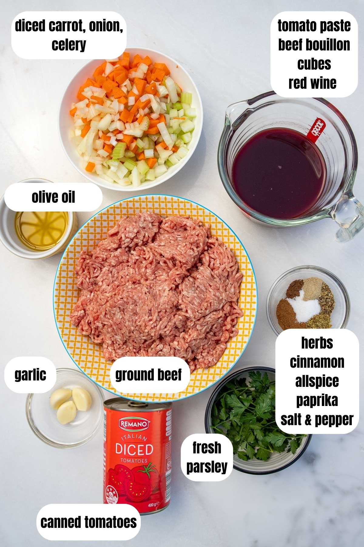 Overhead on marble background of all ingredients for a Greek ragu sauce for pastitsio including ground beef, garlic, herbs and spices, canned, tomatoes, wine, tomato paste and beef bouillon cubes, fresh parsley, olive oil and diced carrot, celery and onion.