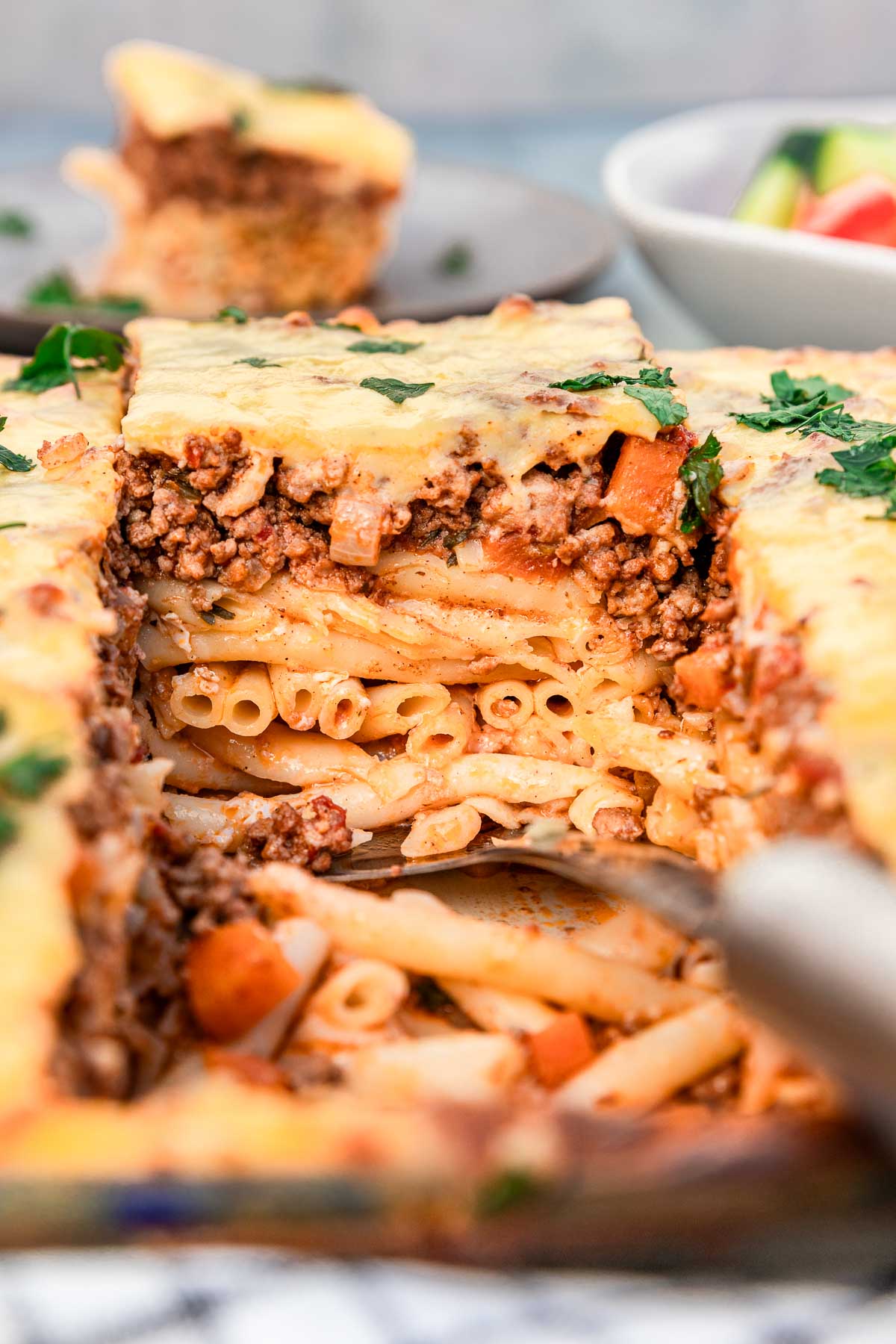 Someone lifting out a square of Greek pastitsio with 3 layers - tube pasta, beef ragu sauce and bechamel sauce, from a large baking dish with a Greek salad and a plate of pastitsio just visible in the background.