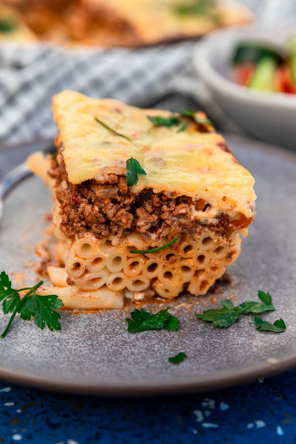 A square of Greek pastitsio with 3 layers - tube pasta, beef ragu sauce and bechamel sauce, on a blue plate and blue background and with a Greek salad and the baking pan with more pastitsio just visible in the background.