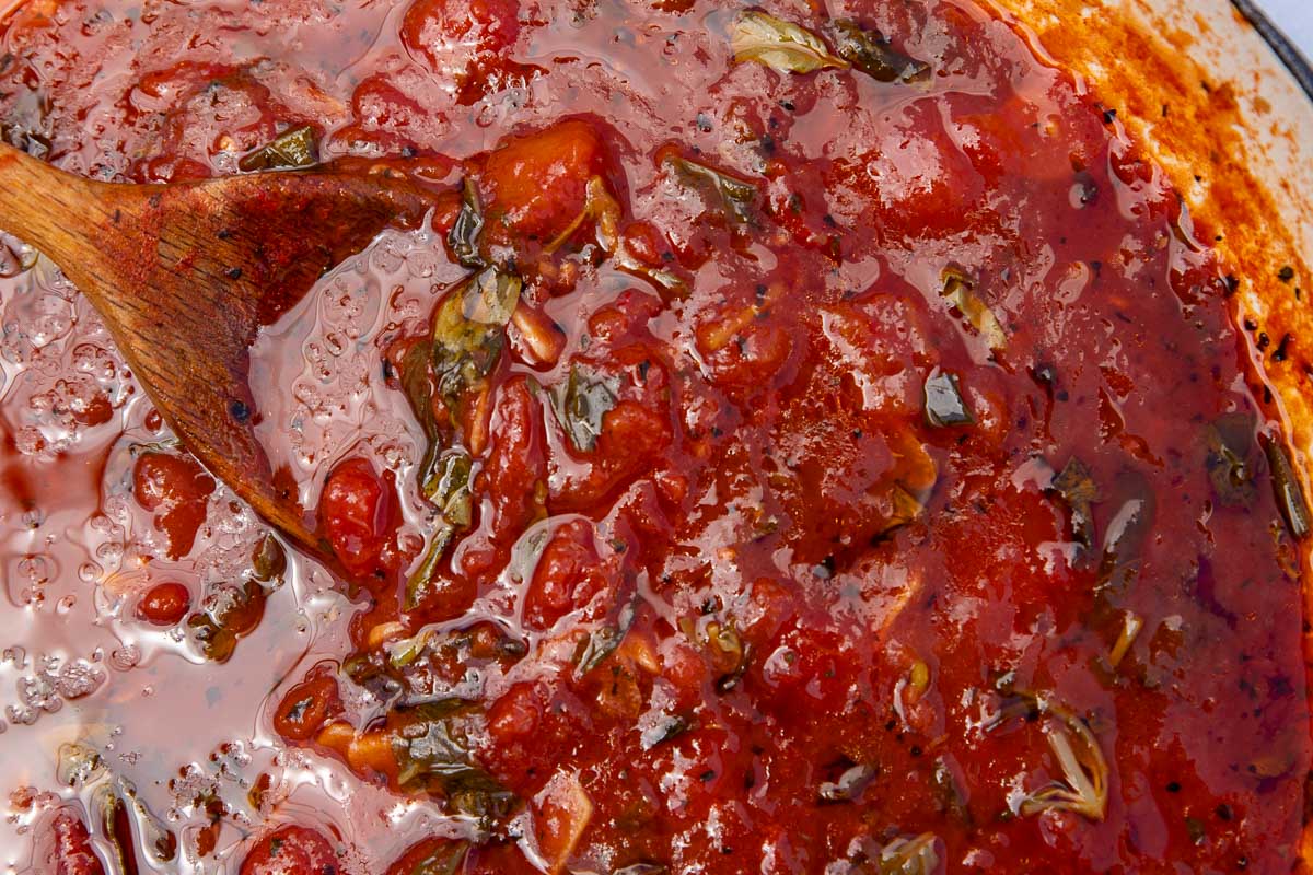 Closeup of the top of a pan of homemade marinara sauce with a wooden spoon in it.