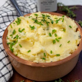 A clay pot of truffle mashed potatoes with chives on top with a spoon in it and fresh thyme at the side.