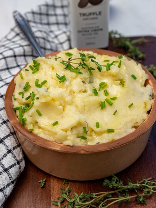 A clay pot of truffle mashed potatoes with chives on top with a spoon in it and fresh thyme at the side.