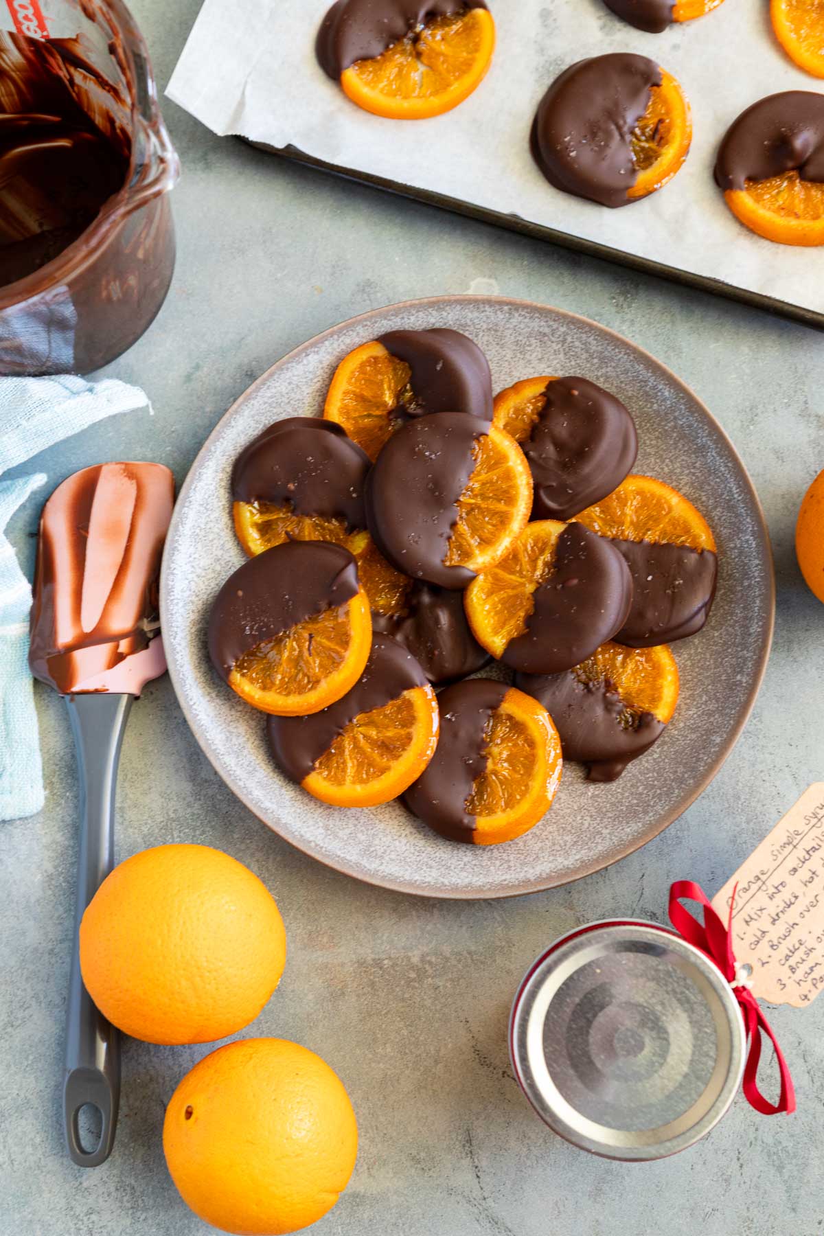 A mottled blue plater of candied orange slices dipped in chocolate with oranges and kitchen equipment around them.