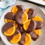 A mottled blue plater of candied orange slices dipped in chocolate with oranges and kitchen equipment around them.