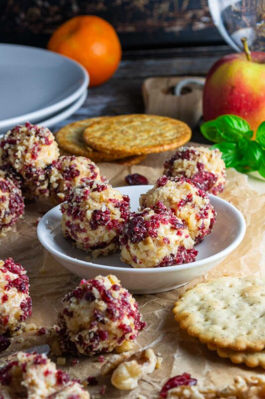 Mini cranberry cheese balls covered in tiny cranberry and walnut pieces on brown paper with crackers and ingredients in the background.