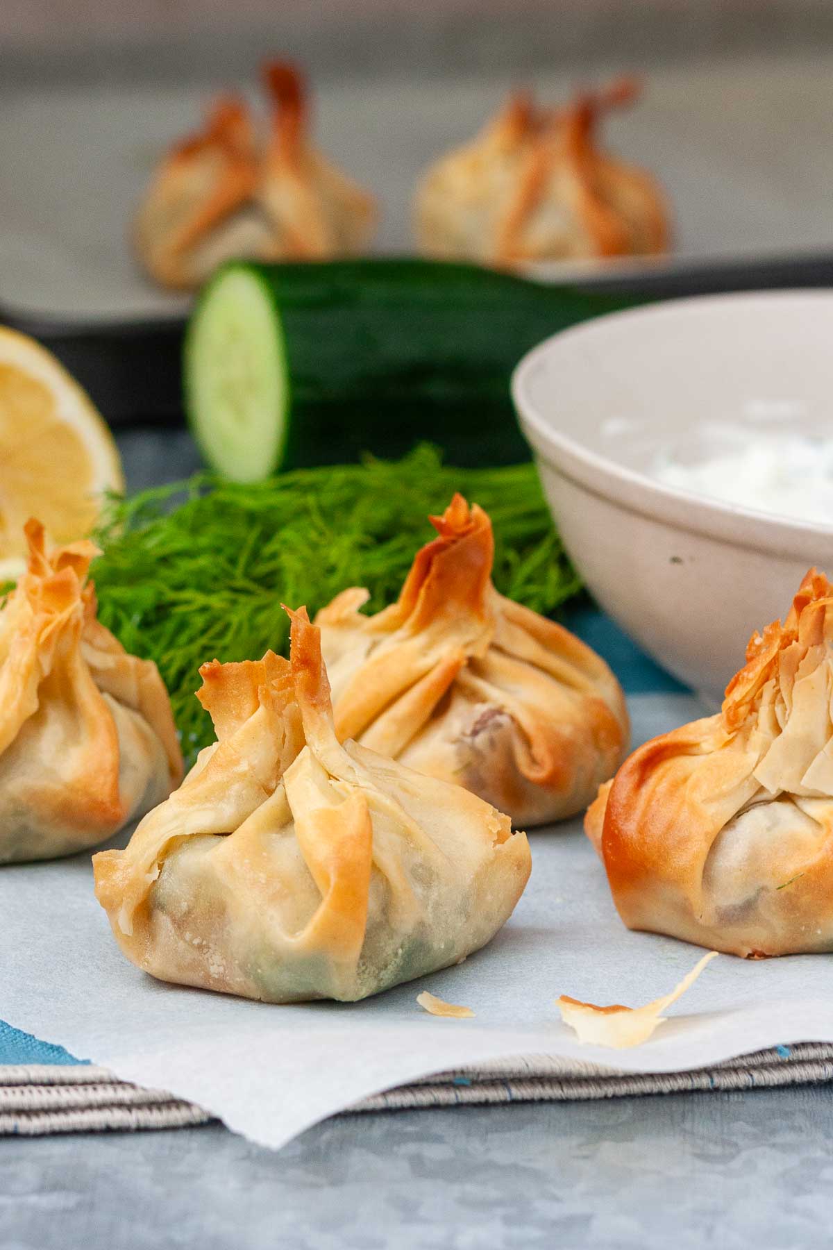 Crispy spinach and feta filo parcels on baking paper and a blue background with some ingredients in the background.