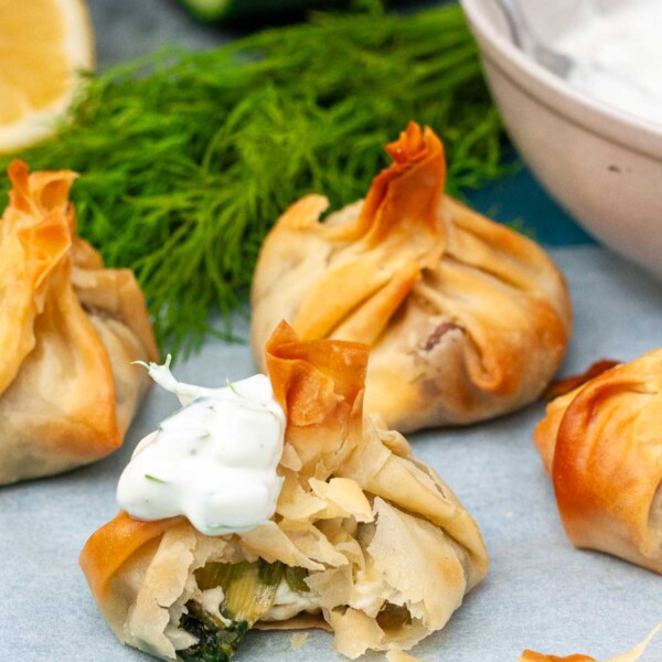 Crispy spinach and feta filo parcels, one with a bite out of it to show the spinach and feta filling inside, all on baking paper with ingredients behind.