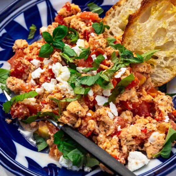 Greek scrambled eggs with tomatoes and crumbled feta and fresh herbs in a blue patterned bowl with a black fork and bread tucked into the dish.
