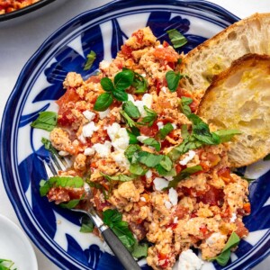 Greek scrambled eggs with tomatoes and feta in a blue patterned bowl with a black fork in it and bread.