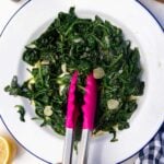 Someone's hand grabbing some sauteed frozen spinach with sliced garlic, lemon and olive oil with pink kitchen tongs with a blue and white tea towel next to the plate.