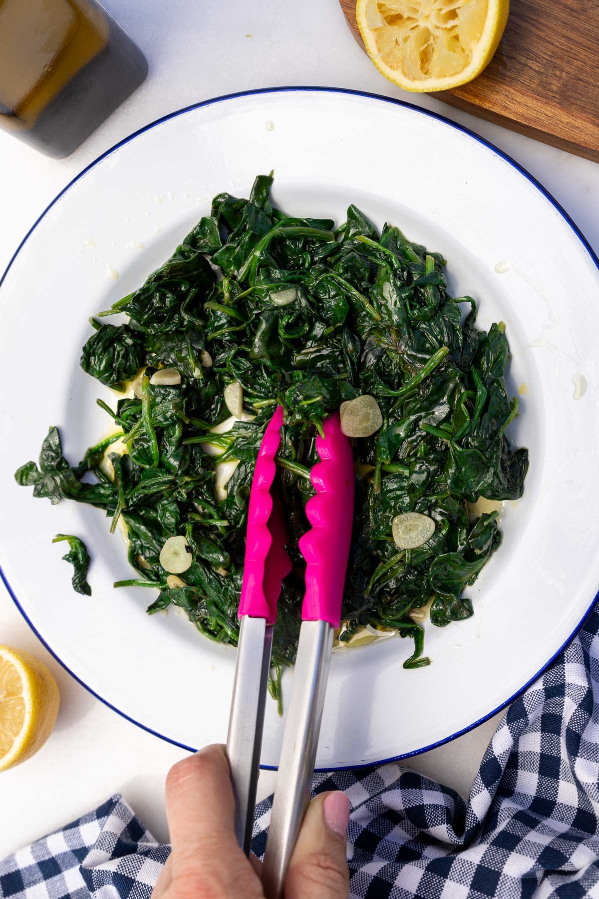 Someone's hand grabbing some sauteed frozen spinach with sliced garlic, lemon and olive oil with pink kitchen tongs with a blue and white tea towel next to the plate.