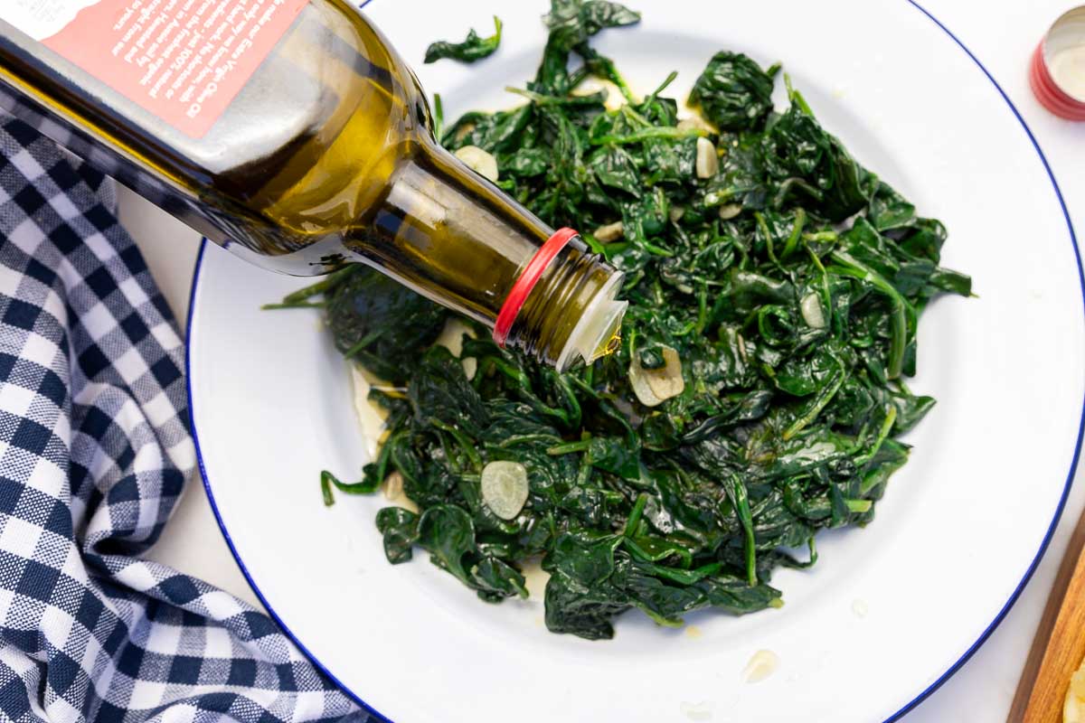 Someone pouring olive oil from a bottle over a white plate of sautéed frozen spinach with garlic slices.