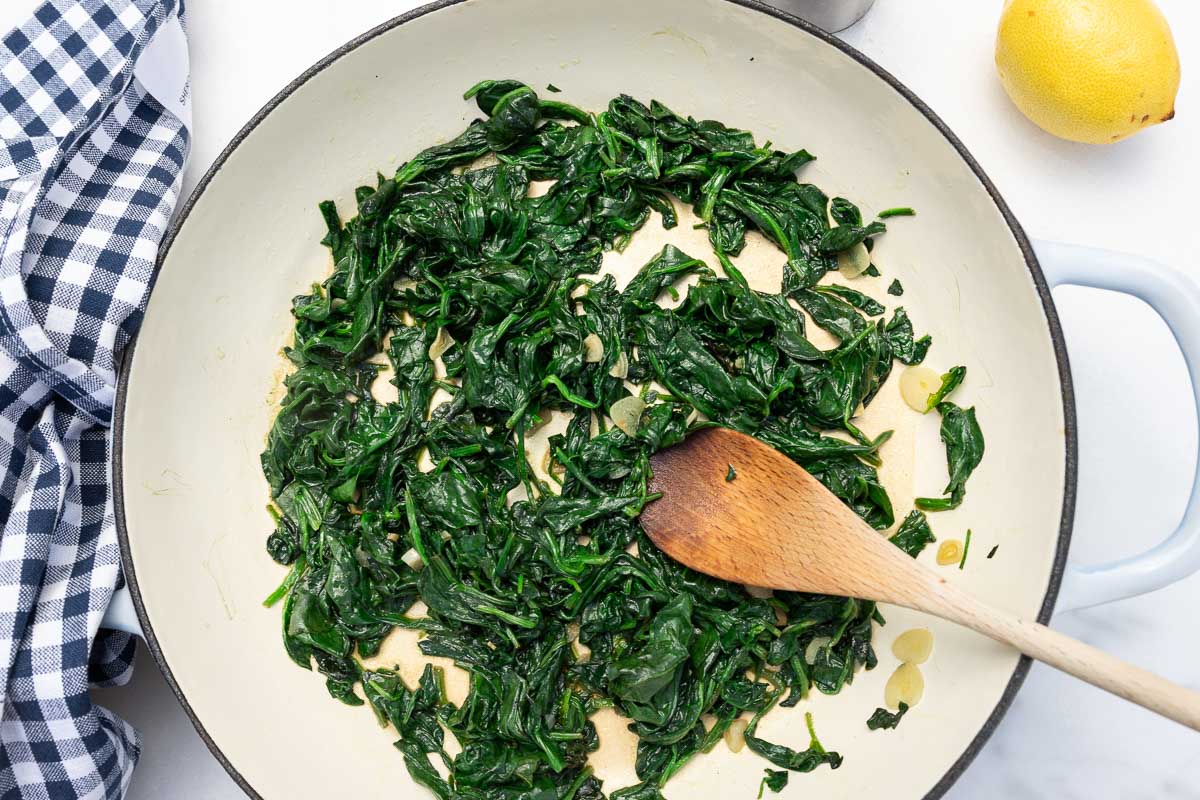 Sautéed frozen spinach and garlic in a cast iron pan with wooden spoon and with a blue and white checked tea towel at the side.