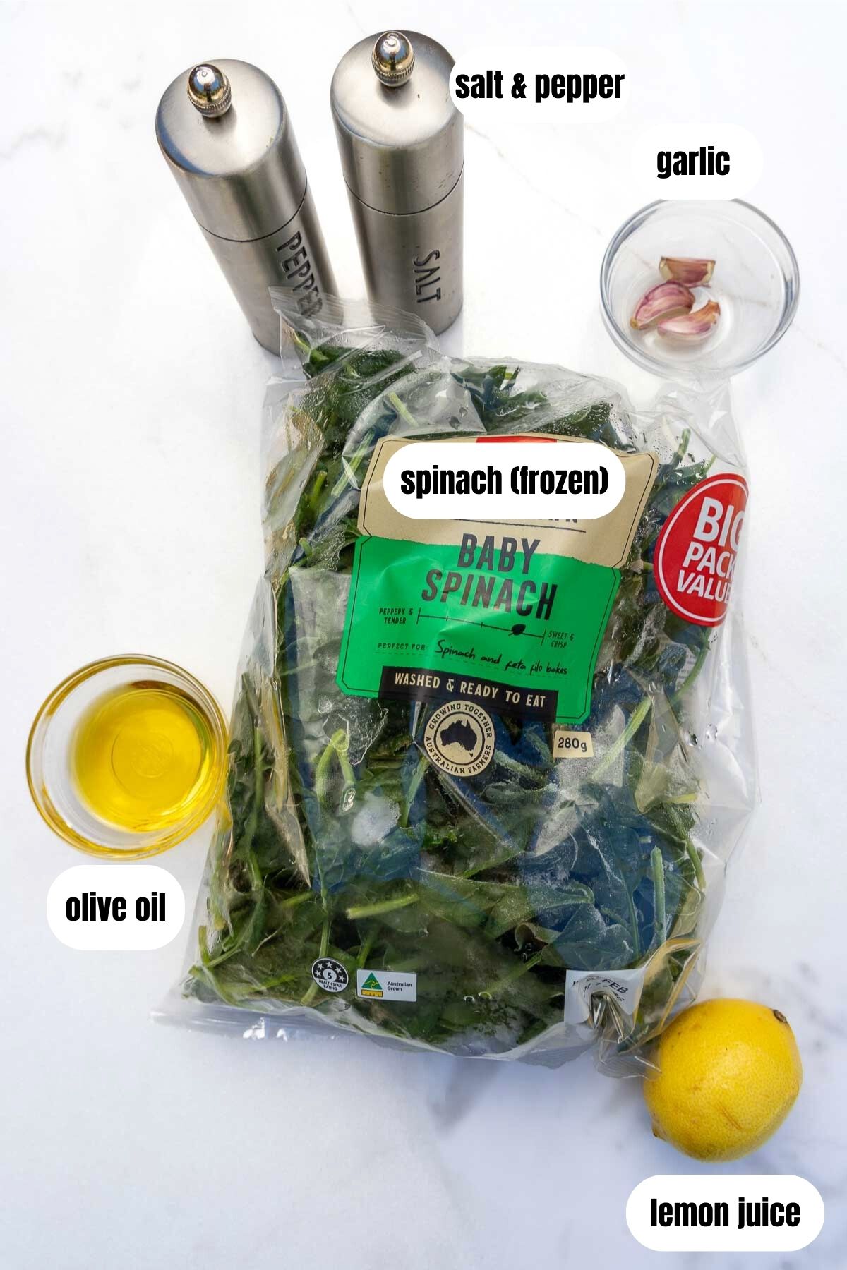 All ingredients for sautéed frozen spinach on a marble background including a big bag of spinach, garlic cloves, a lemon, olive oil and salt and pepper.
