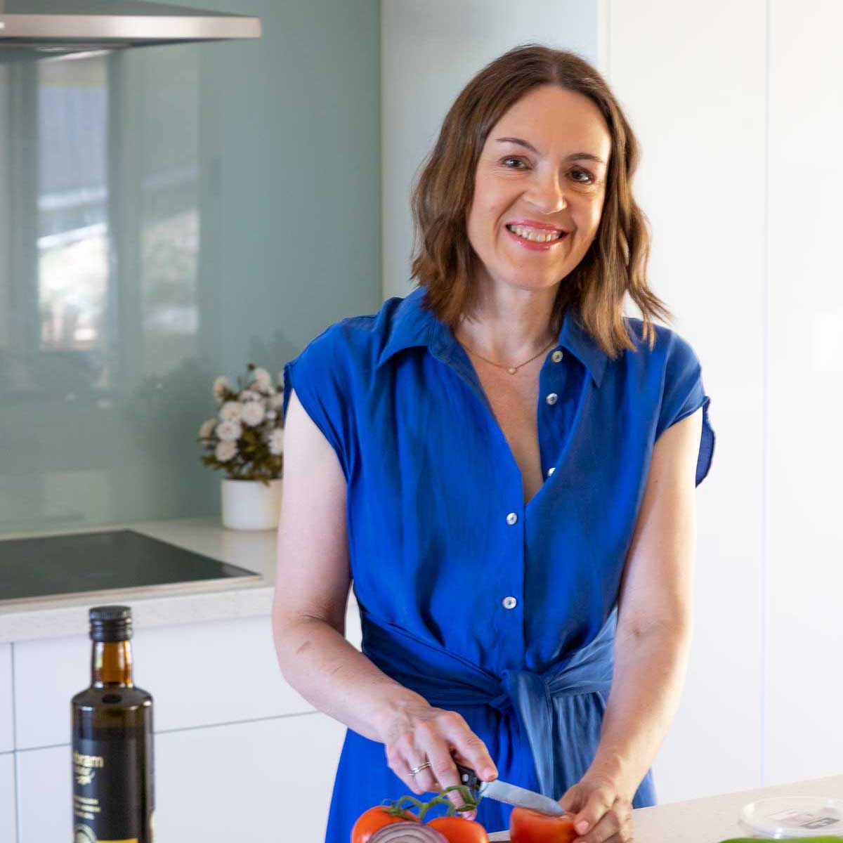 A square image of Helen Schofield, owner of food blog Scrummy Lane, standing at her bench in the kitchen smiling and wearing a blue dress and chopping tomatoes.