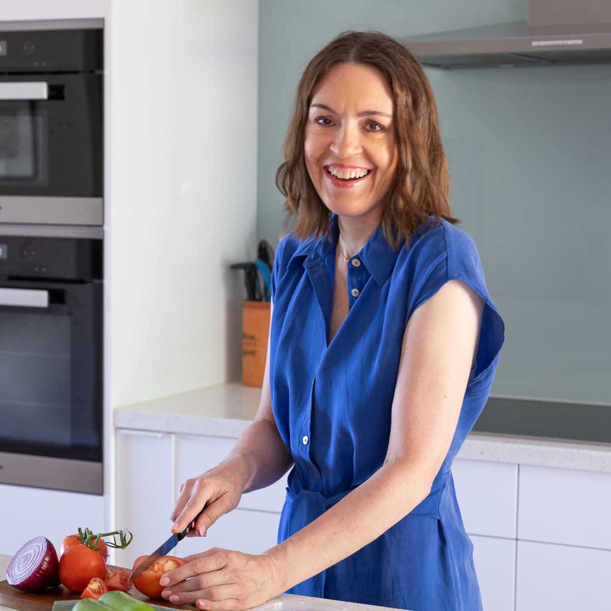 A square image of Helen Schofield, owner of food blog Scrummy Lane, standing at her bench in the kitchen smiling and wearing a blue dress and chopping vegetables.
