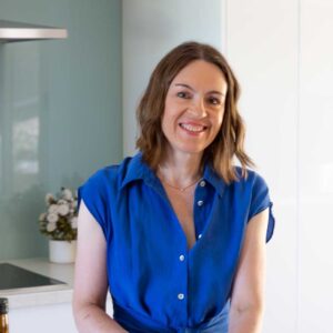A square image of Helen Schofield, owner of food blog Scrummy Lane, standing at her bench in the kitchen smiling and wearing a blue dress and chopping vegetables for a salad.