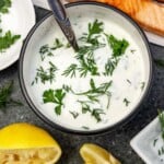 An overhead view of a small off-white bowl of Greek yogurt dill sauce on a dark grey background with lemon segments and cooked salmon visible at the side