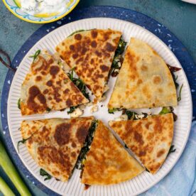 Greek quesadilla triangles arranged in a circle on a white plate with blue tea towel and ingredients around it.