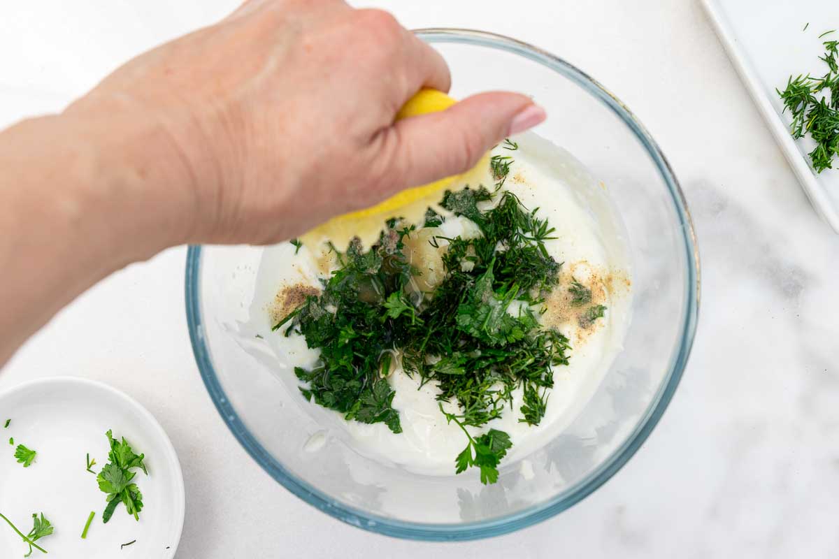 Someone's hand squeezing half a lemon into a glass bowl of Greek yogurt, fresh herbs, garlic and salt and pepper.