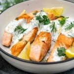 Closeup of a dish of cooked salmon with dill yogurt sauce drizzled over it garnished with lemon segments and herbs and with a green and cream tea towel at the side.