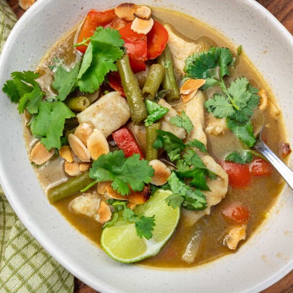 A portion of Thai fish curry and a spoon in a light beige bowl on a wooden board with a green tea towel at the side.